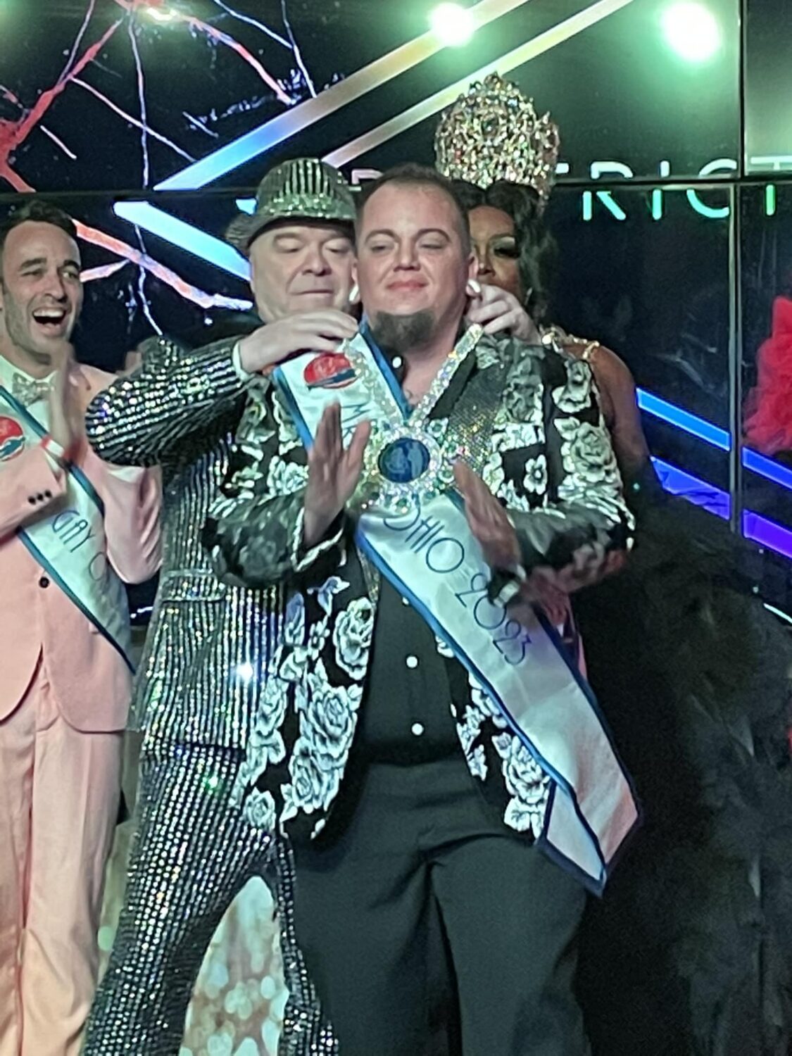 Joey Fleming medals Ty Erup as the new Mr. Gay Ohio.  Former Mr. Gay Ohio, Matthew Allen Meade (far back left), cheers in the background.  | Mr. and Miss Gay Ohio | District West (Columbus, Ohio) | 9/17-9/18/2022
